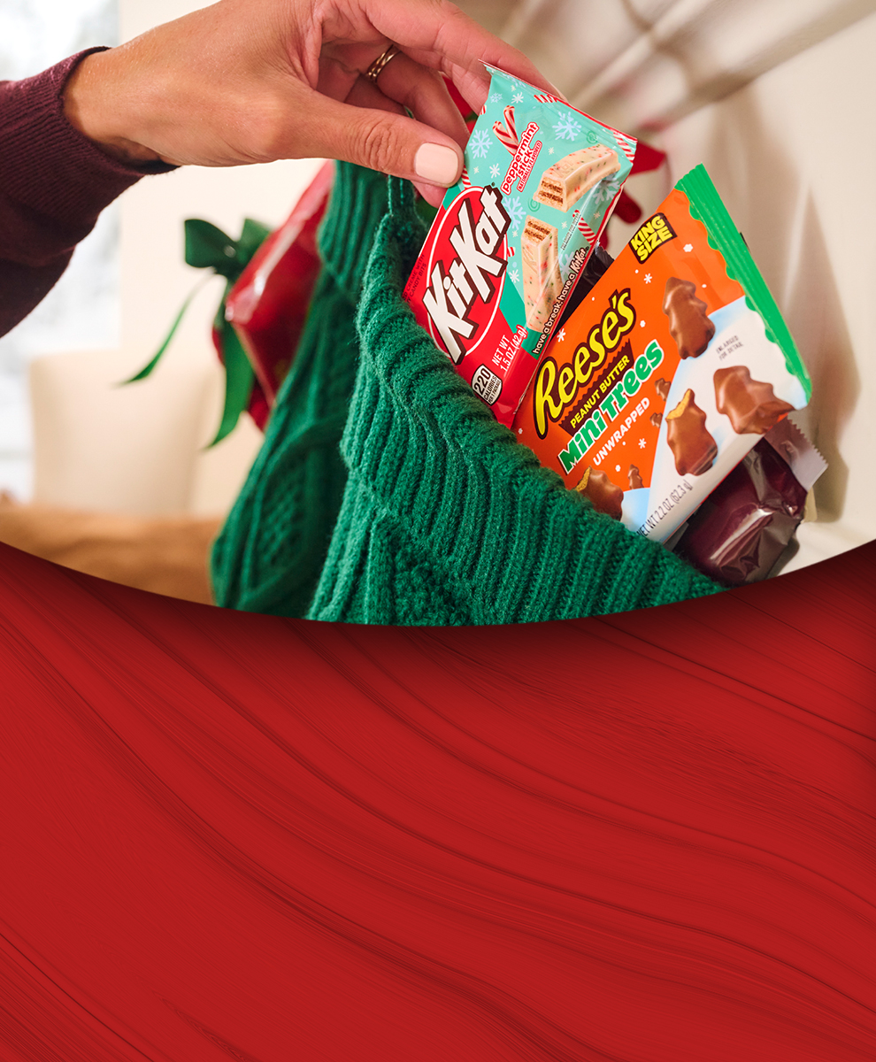 Promotional graphic featuring REESE'S and KIT KAT® assorted candy bags being dropped into a Christmas stocking hanging on a mantel