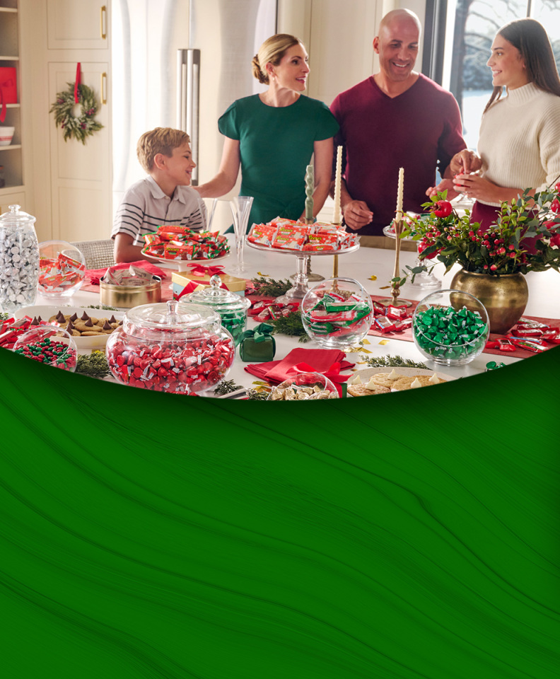 Promotional graphic featuring a family standing around a kitchen island with various bowls and dishes filled with assorted holiday candies