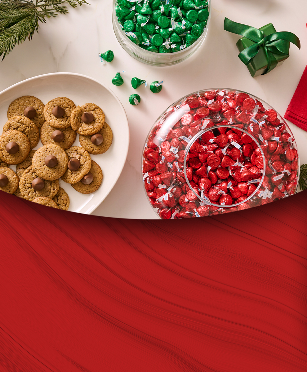 Promotional graphic showing a holiday-themed tabletop spread with wrapped HERSHEY'S Kisses in a glass bowl and HERSHEY'S Kisses-topped freshly baked cookies