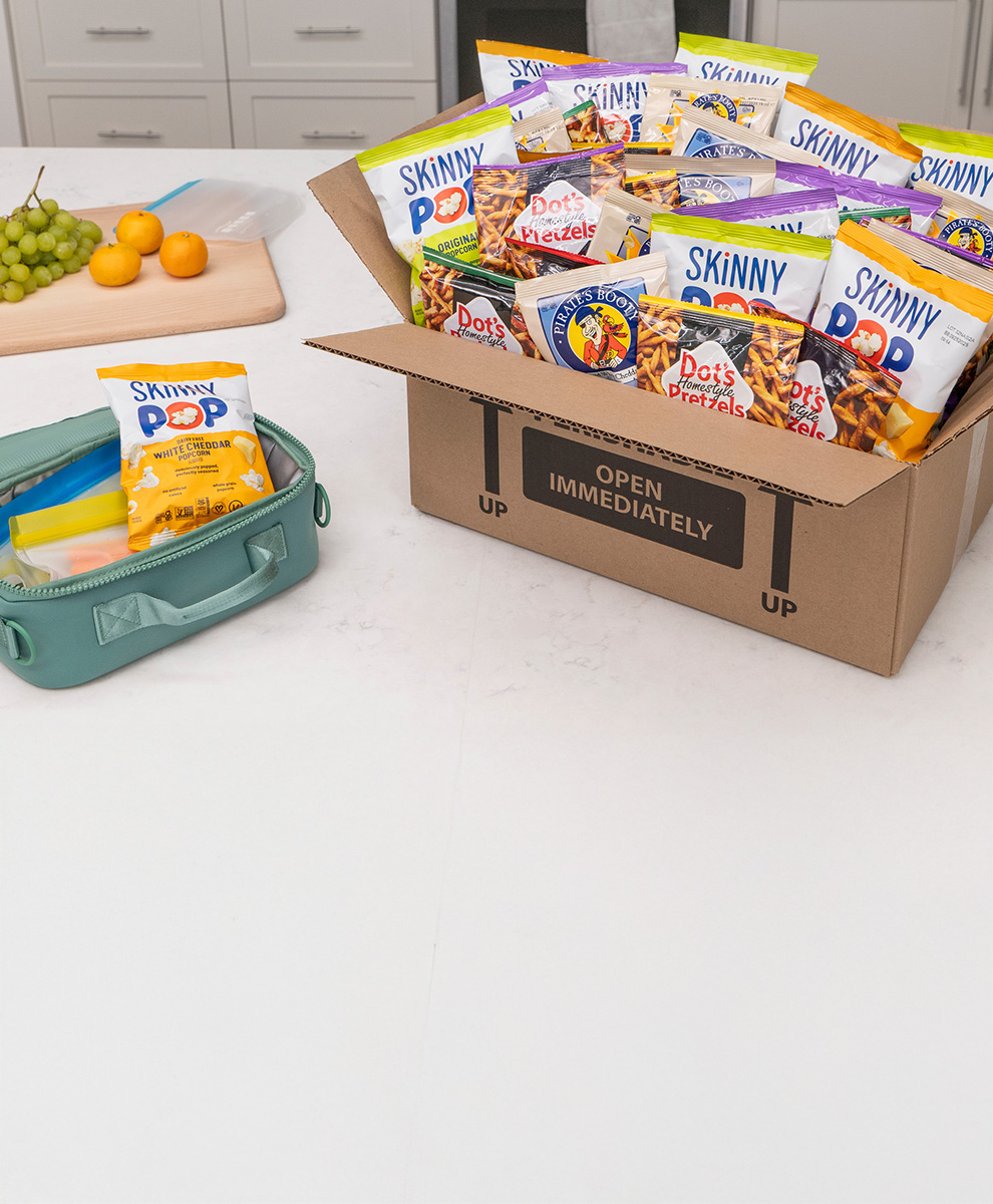 Promotional graphic showing a Pick Your Pack cardboard box filled with individually-sized bags of snacks on a countertop next to a lunchbox being packed