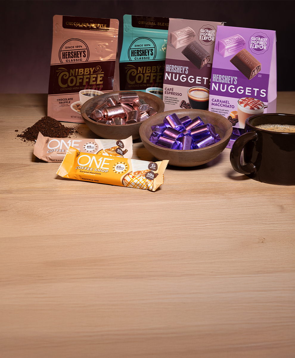 HERSHEY'S Cafe Espresso and Caramel Macchiato Chocolate Nuggets and Nibby Coffee and ONE Coffeeshop Protein Bars displayed on a wooden tabletop