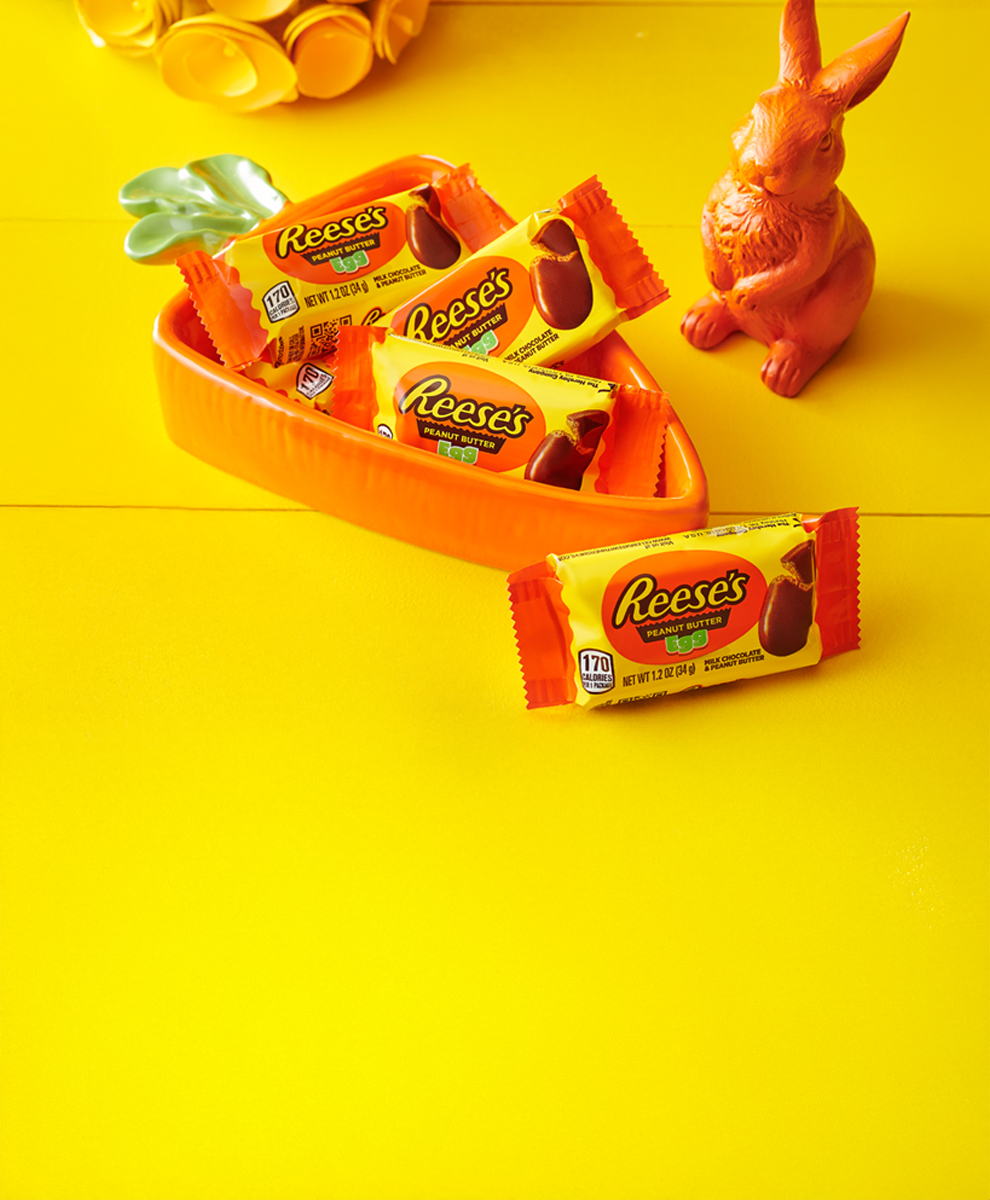 Individually wrapped REESE'S Peanut Butter Eggs in a carrot-shaped bowl on a yellow wooden tabletop