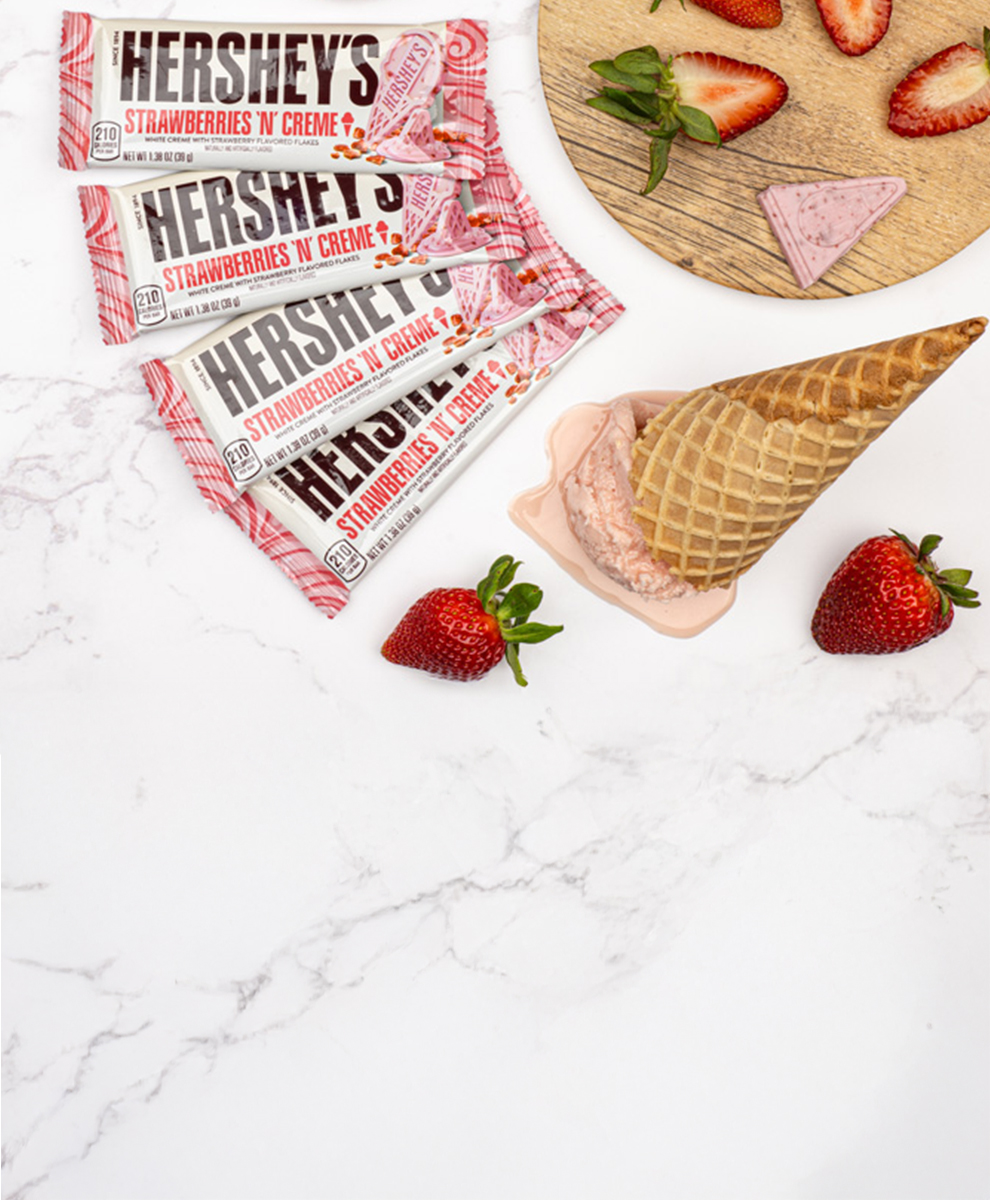 Promotional graphic featuring HERSHEY's Strawberries N Creme White Chocolate Bar melting on a marble table with cut up strawberries
