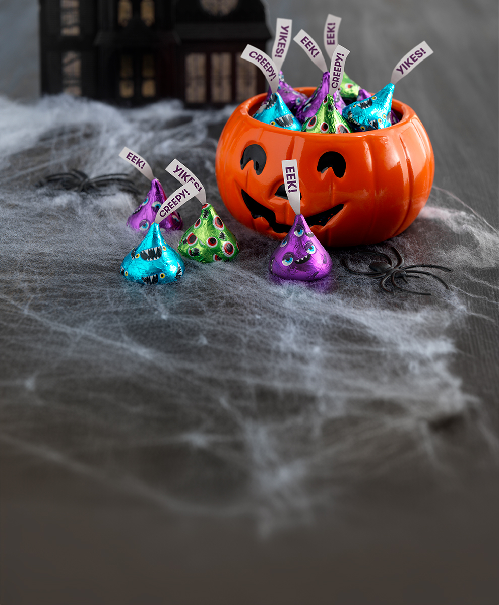 Promotional graphic featuring HERSHEY'S Kisses in monster-themed foils in a pumpkin-shaped bowl