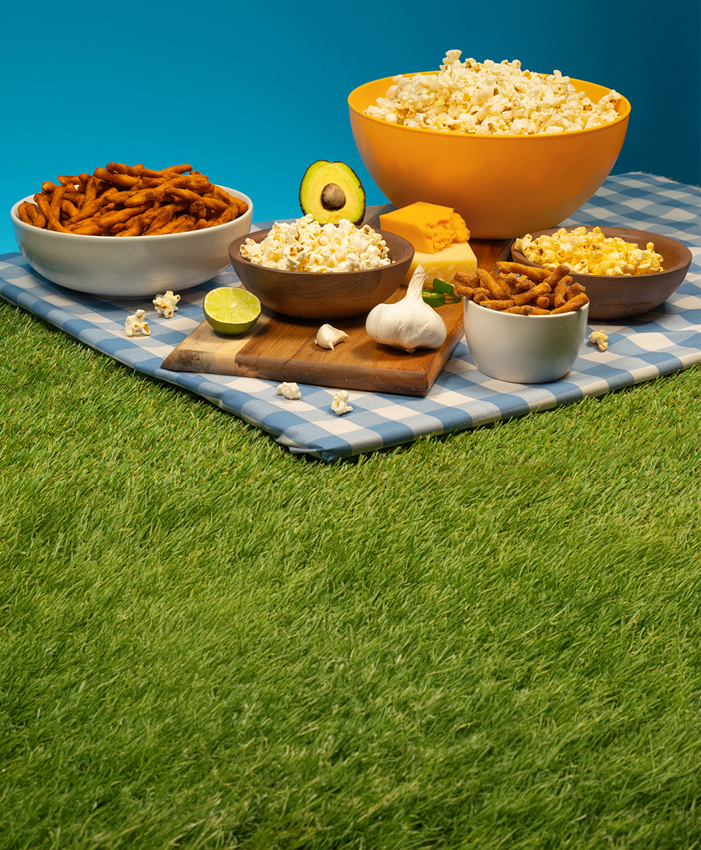 Salty snacks including SKINNYPOP popcorn and DOT'S Pretzel Sticks in assorted bowls set up on a picnic blanket
