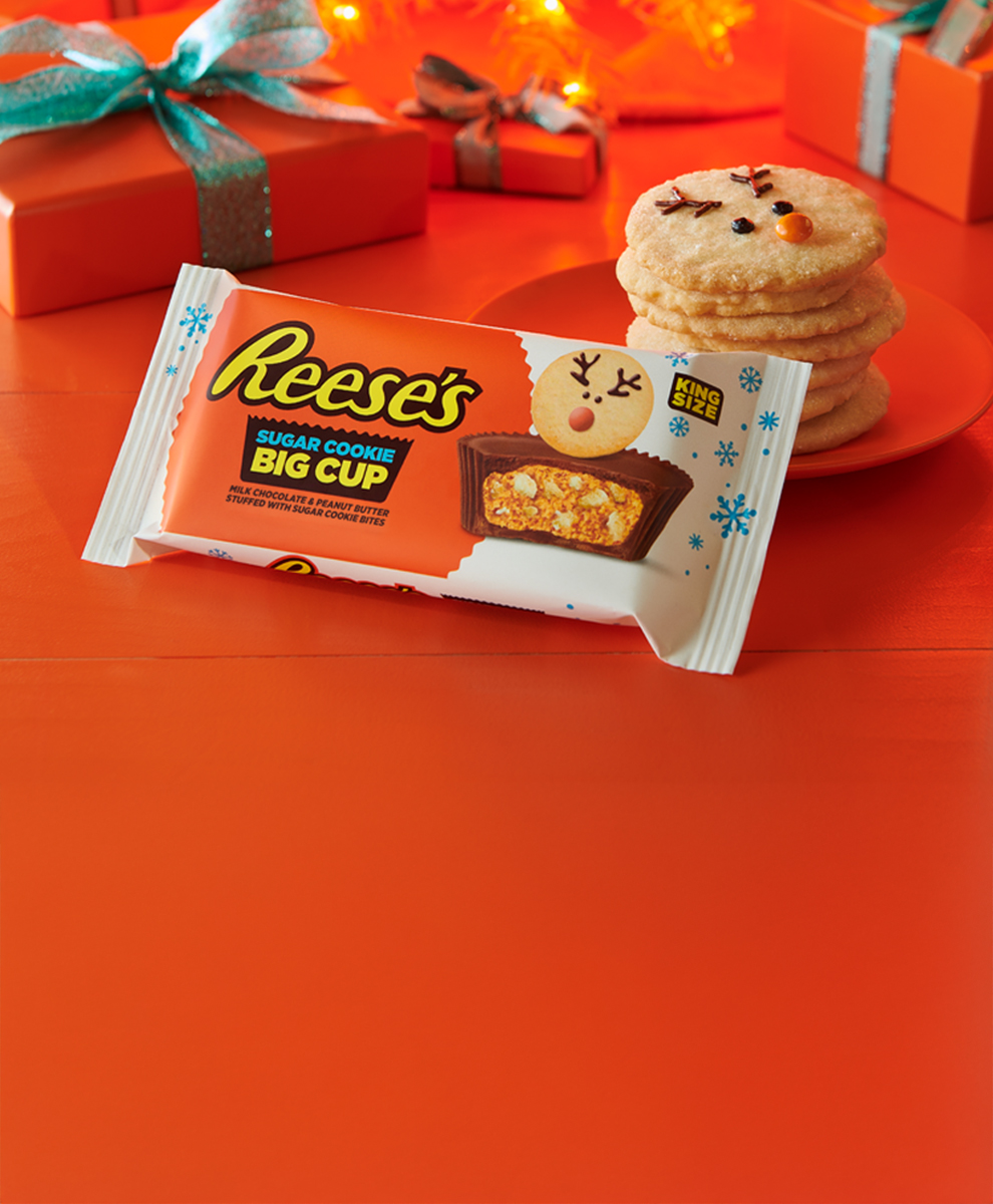 REESE'S peanut butter cups with sugar cookie pieces candy pack on an orange background placed near wrapped gifts and plates of cookies