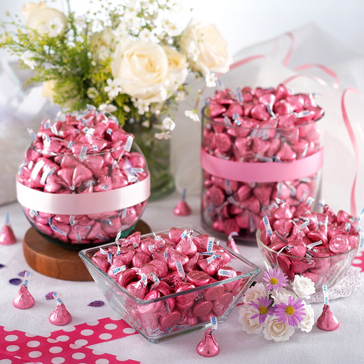 HERSHEY'S Kisses in pink foils filling various sized and shaped glass jars on a tablecloth-covered table
