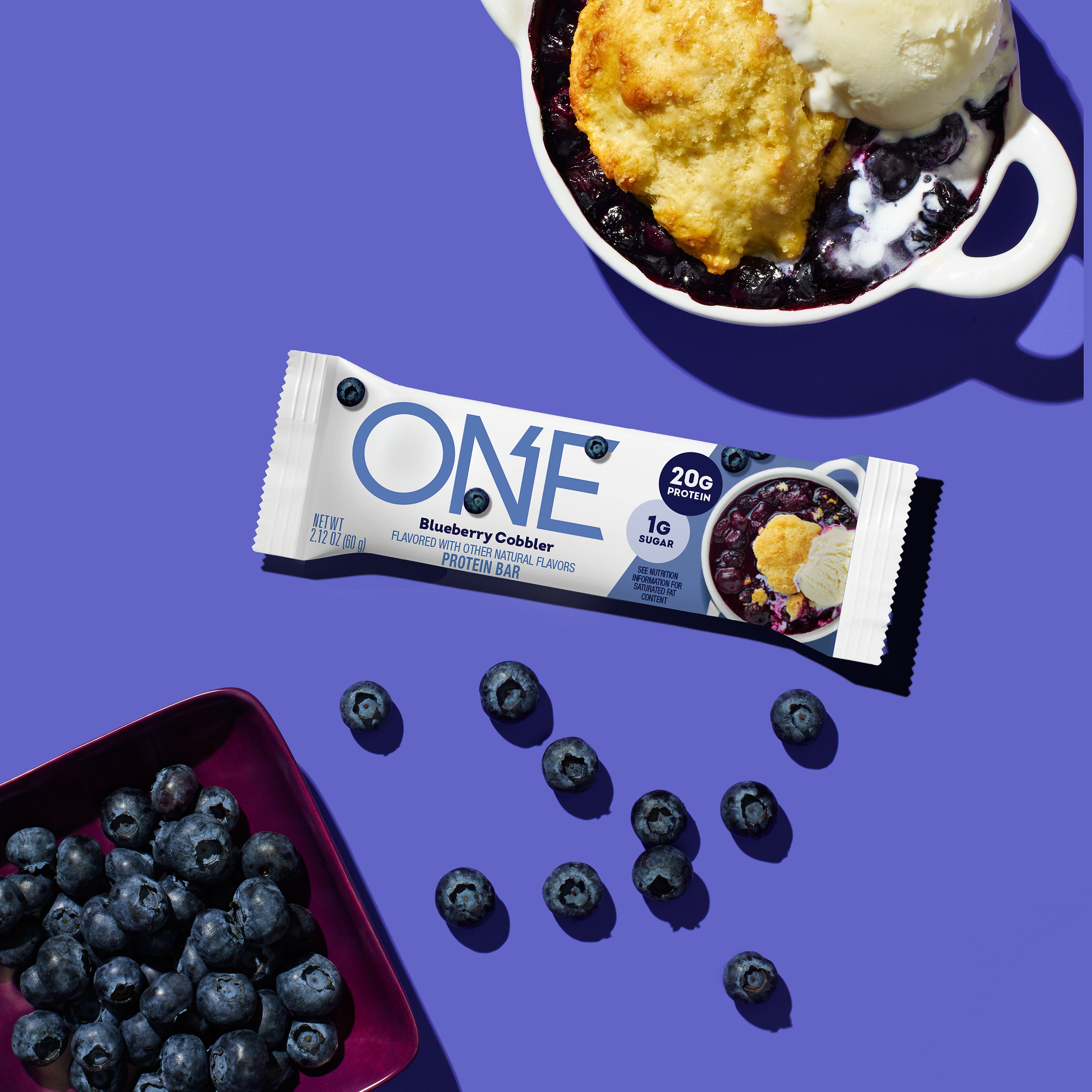 ONE Bar Blueberry Cobbler Flavored Protein Bars displayed next to a tray of blueberries and blueberry cobbler in a dutch oven