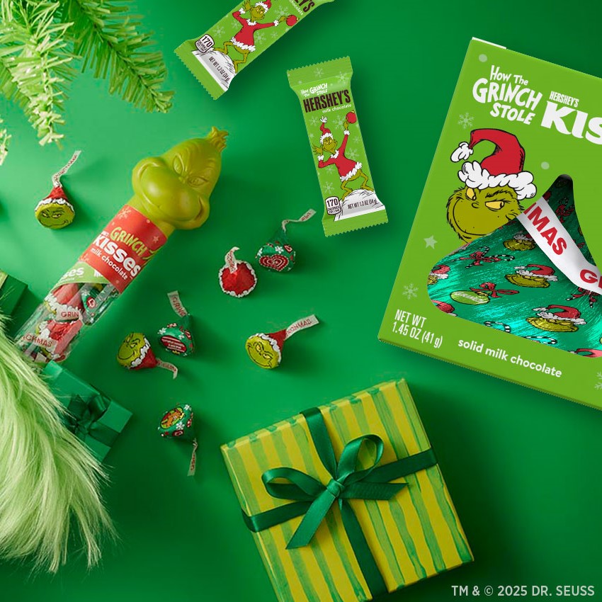 Promotional graphic showing HERSHEY'S Grinch seasonal collection displayed on a green background arranged next to wrapped presents and holiday-themed greenery