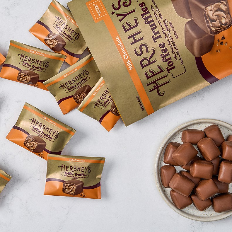 Promotional graphic showing HERSHEY'S Grinch seasonal collection displayed on a green background arranged next to wrapped presents and holiday-themed greenery