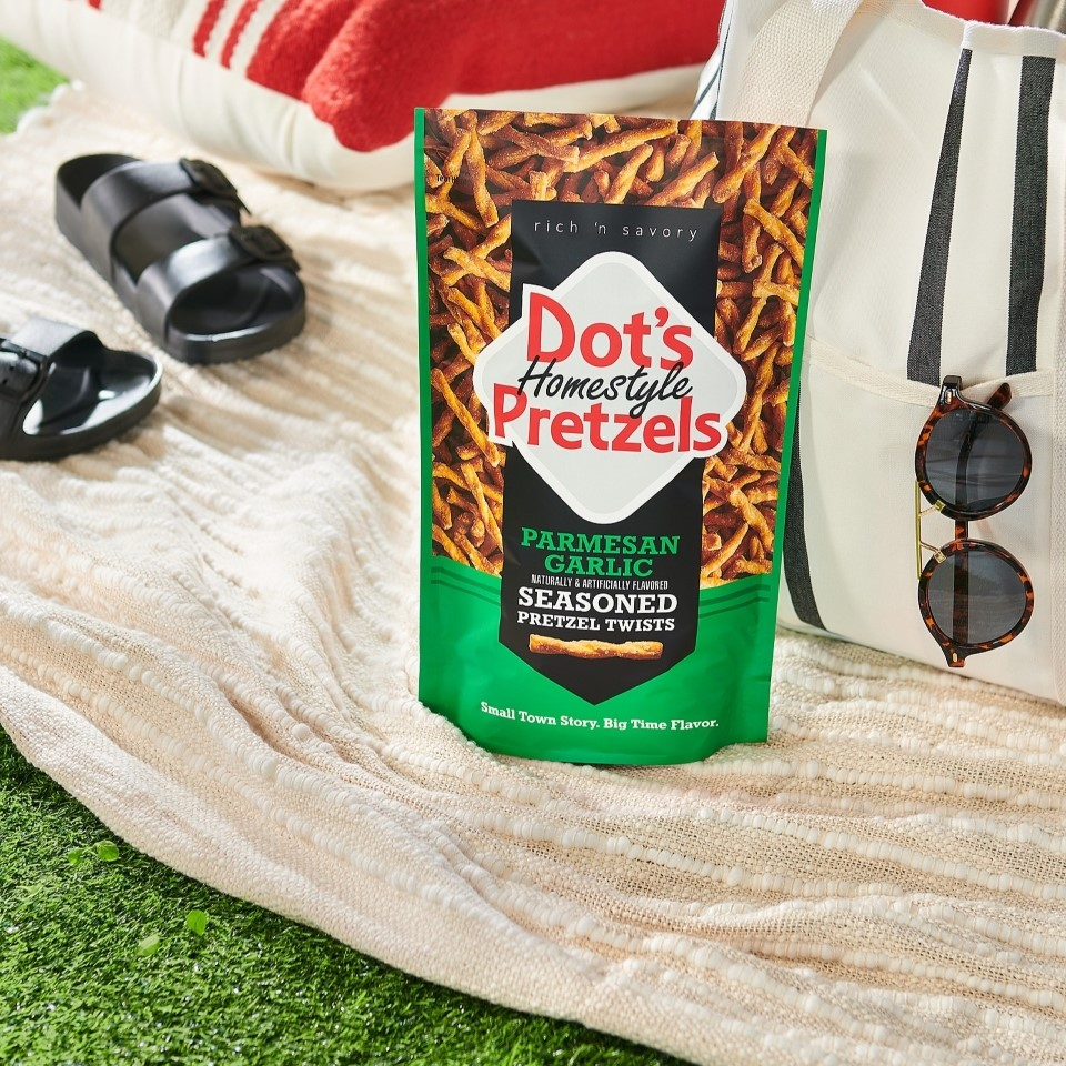 DOT'S Garlic Parmesan Flavored Pretzels Twists bag on a blanket laid over grass alongside a tote bag and pair of sandals
