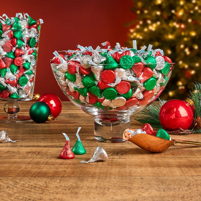Bulk HERSHEY'S KISSES in red and green and silver foils displayed in various glass bowls on a wooden tabletop