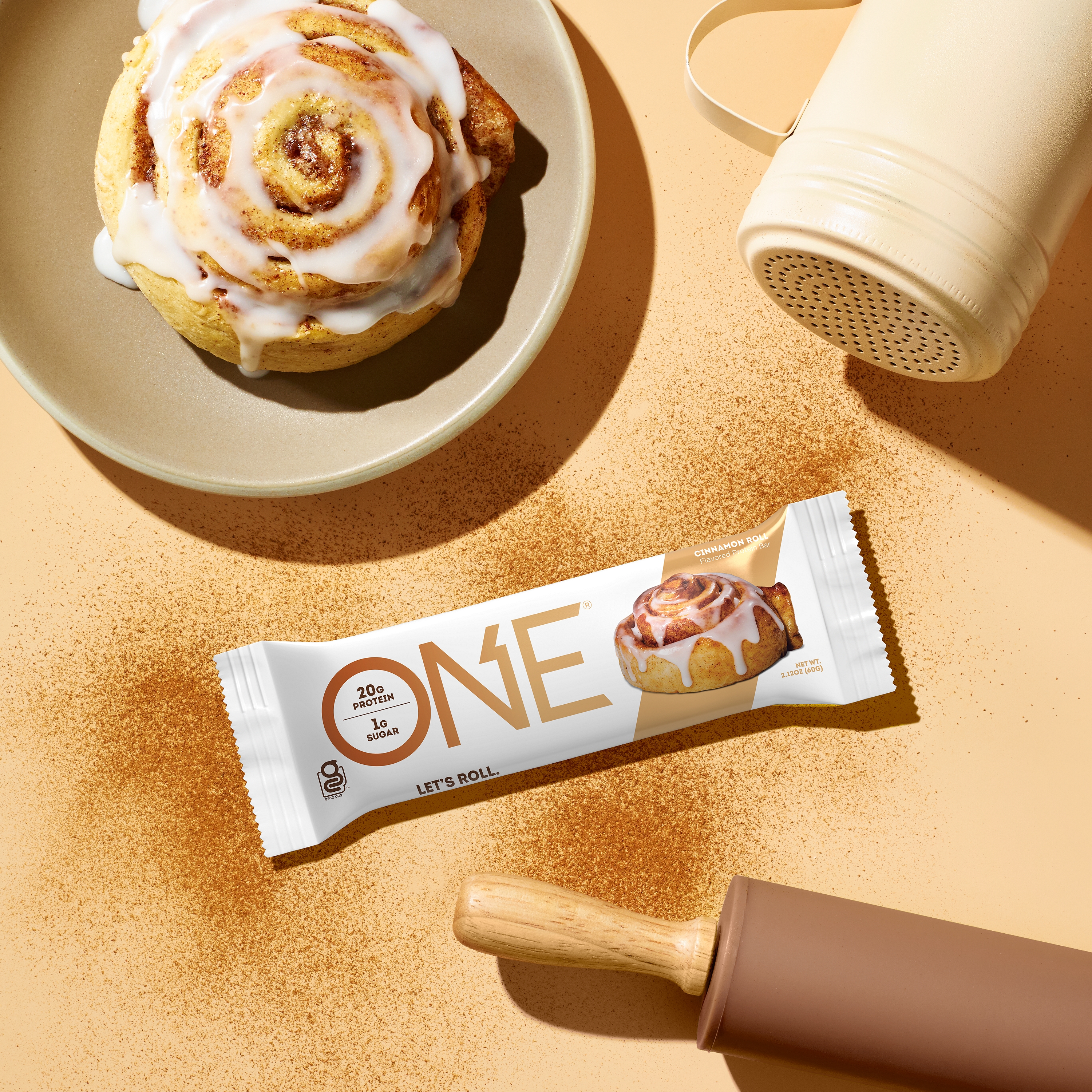 Promotional graphic featuring a Cinnamon Roll flavored ONE Brand protein bar arranged next to a cinnamon roll and baking tools